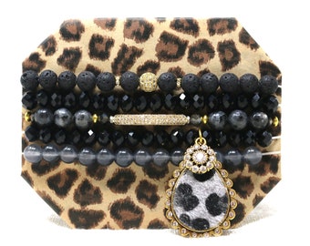 May include: A set of five bracelets featuring black lava beads, black crystal beads, grey gemstone beads, and a gold charm with a leopard print design. The bracelets are displayed on a leopard print background.
