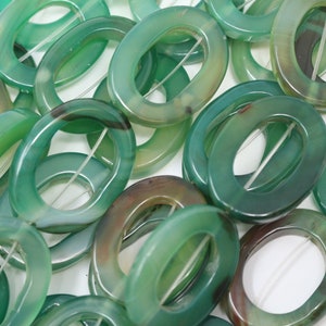 Könnte beinhalten: Nahaufnahme eines Stapels grüner Achat-Ovalperlen mit einem Loch in der Mitte. Die Perlen sind glatt und poliert und haben ein natürliches, organisches Aussehen.