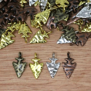 Puede incluir: Una colección de dijes de metal con forma de punta de flecha en varios acabados, incluyendo oro brillante, plata, bronce antiguo y cobre. Cada dije presenta una superficie texturizada y un pequeño lazo en la parte superior para su fijación, dispuestos en un montón con cuatro piezas individuales colocadas en una fila en la parte inferior.