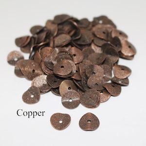 May include: A pile of copper-colored metal heart-shaped beads with a hole in the center. The beads are scattered on a white surface. The word "Copper" is written below the beads.