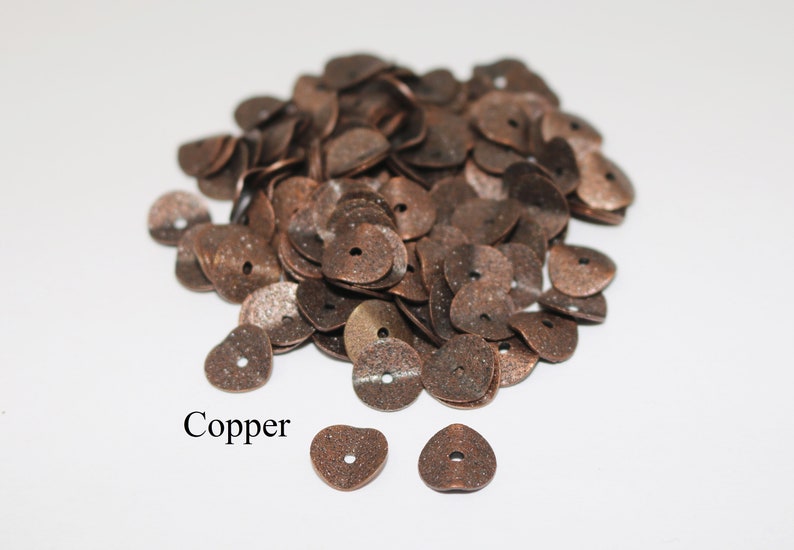 May include: A pile of copper-colored metal heart-shaped beads with a hole in the center. The beads are scattered on a white surface. The word "Copper" is written below the beads.