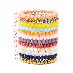 May include: A stack of 12 colorful beaded bracelets, each with a different color and style. The bracelets are stacked in order from top to bottom, with the top bracelet being peach, followed by iridescent, yellow, light blue, orange, pink, purple, red, beige, clear, dark blue, and gold.