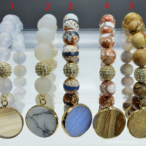 Op de afbeelding: Vijf armbanden met verschillende gekleurde kralen en gouden accenten. Elke armband heeft een ronde hanger met een andere steen. De hangers zijn gemaakt van agaat, wit marmer, blauw agaat en bruin agaat.