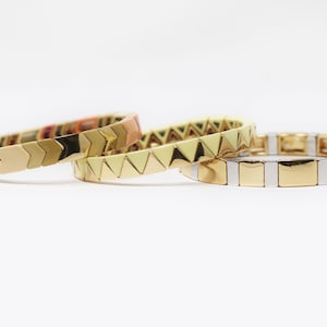May include: Three gold bracelets with different coloured enamel designs. The bracelets are stacked on top of each other. The top bracelet has a light yellow enamel design, the middle bracelet has a light green enamel design, and the bottom bracelet has a light pink enamel design.