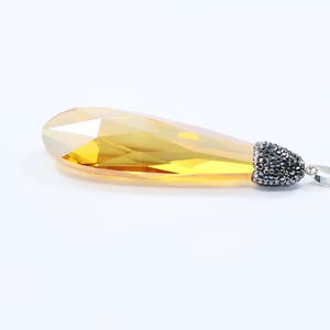May include: A teardrop-shaped pendant with a faceted, yellow crystal and a silver bail adorned with black rhinestones.
