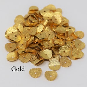 May include: A pile of gold-colored metal heart-shaped sequins with a hole in the center.  The sequins have a textured surface.  Gold.