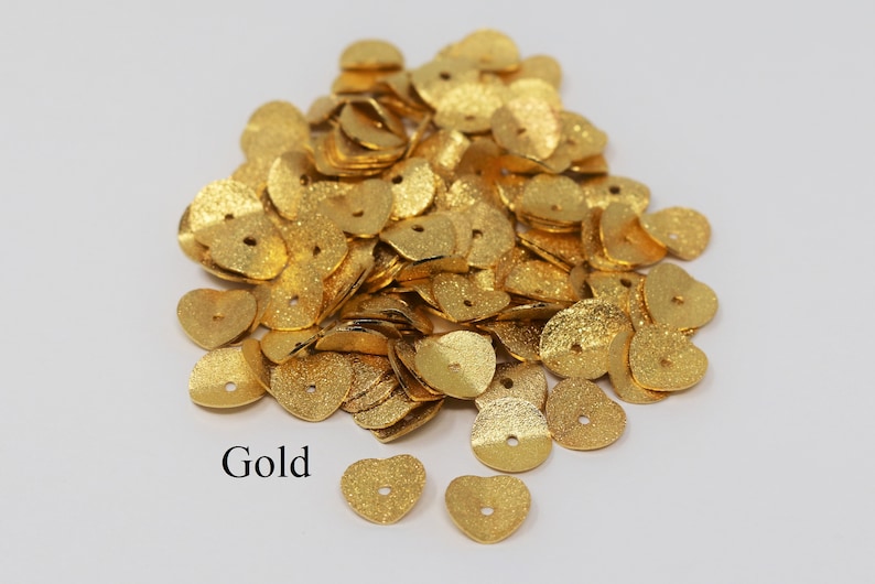 May include: A pile of gold-colored metal heart-shaped sequins with a hole in the center.  The sequins have a textured surface.  Gold.