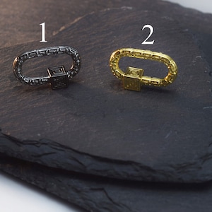 May include: Two metal clasps, one black and one gold, with a square design in the center. The clasps are on a black slate surface.