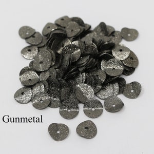 May include: A pile of gunmetal-colored sequins with a hole in the center. The sequins are scattered on a white surface. Gunmetal is written below the sequins.