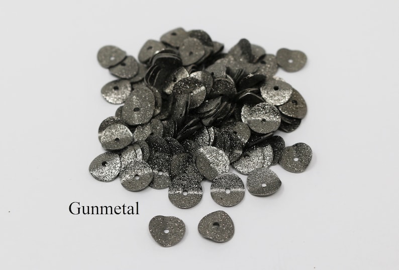 May include: A pile of gunmetal-colored sequins with a hole in the center. The sequins are scattered on a white surface. Gunmetal is written below the sequins.