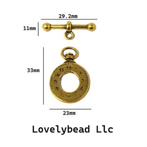 Puede incluir: Un cierre de palanca en tono latón que incluye una barra de 29,2 mm por 11 mm y un anillo circular con números de reloj que mide 33 mm por 23 mm. El texto Lovelybead Llc aparece impreso en la parte inferior de la imagen.
