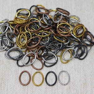 Puede incluir: Un conjunto de anillas ovaladas en varios acabados metálicos, incluyendo oro, plata, cobre y negro. Las anillas están dispersas sobre una superficie de tela de color neutro, con algunas anillas cuidadosamente dispuestas.