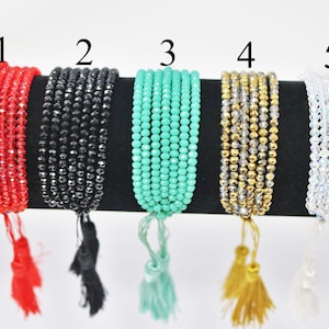 Könnte beinhalten: Fünf Armbänder mit unterschiedlich farbigen Perlen und Quasten. Die Armbänder sind von 1 bis 5 nummeriert. Die Perlen sind rot, schwarz, grün, gold und klar.
