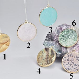 May include: Six round pendants featuring a variety of stone designs. The pendants are gold-toned and have a hole at the top for hanging. The stones come in various colours and patterns, including brown, white, green, and blue.
