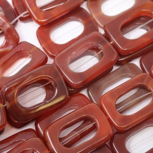 May include: A strand of carnelian beads with a rectangular shape and a hole in the center. The beads are a deep reddish-orange color and have a natural, polished finish.