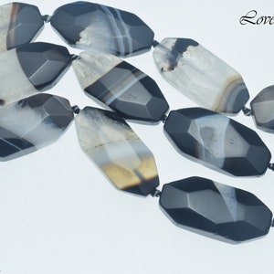 May include: A strand of black and white agate gemstone beads, each bead is faceted and measures 45x20mm. The beads have a natural, organic look with a variety of patterns and textures.