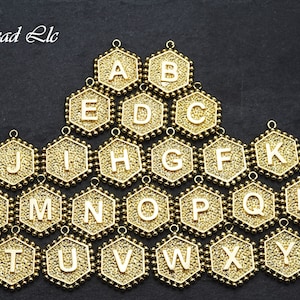 Op de afbeelding: Goudkleurige alfabetletterbedels in de vorm van zeshoeken. Elke bedel heeft een reliëfletter aan de voorkant en een lus aan de bovenkant om te bevestigen aan sieraden of andere knutselwerkjes.