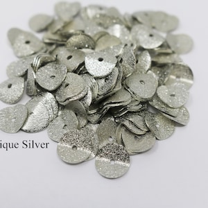 May include: A pile of antique silver heart-shaped metal sequins with a textured finish. The sequins have a small hole in the center.