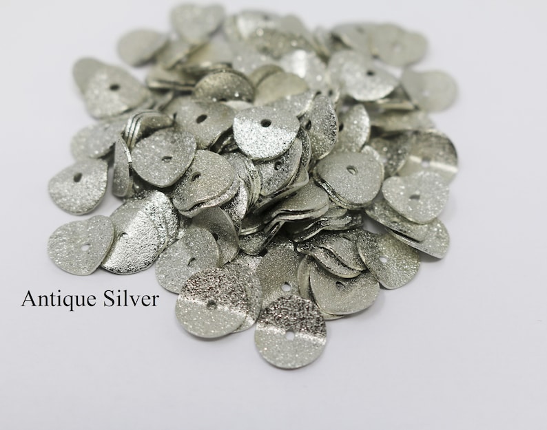 May include: A pile of antique silver heart-shaped metal sequins with a textured finish. The sequins have a small hole in the center.