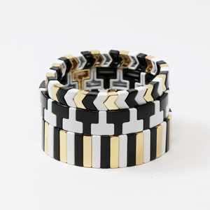 May include: Three stacked bracelets with geometric patterns. The top bracelet has a black and white chevron pattern, the middle bracelet has a black and white cross pattern, and the bottom bracelet has a black, white, and gold stripe pattern.
