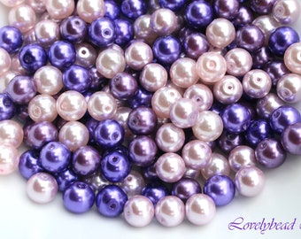 May include: A pile of pearl beads in shades of purple, pink, and white. The beads are round and have a smooth, glossy finish.