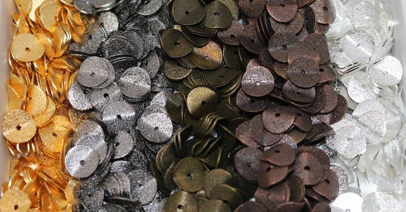 May include: Assortment of metal buttons in various colors, including gold, silver, bronze, and copper. The buttons are all round with a small hole in the center. They are arranged in rows in a container.