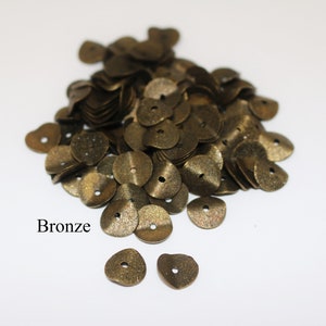 May include: A pile of bronze-colored metal sequins, with two individual sequins in the foreground.  The sequins are shaped like small, flat discs with a hole in the center.