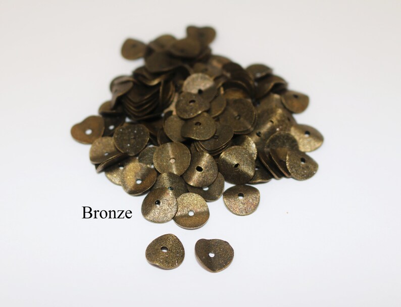 May include: A pile of bronze-colored metal sequins, with two individual sequins in the foreground.  The sequins are shaped like small, flat discs with a hole in the center.