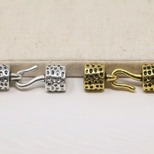 May include: Two metal clasps for jewelry making. One is silver and the other is gold. Both have a textured surface and a hook.