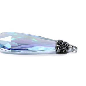 May include: A teardrop-shaped crystal pendant with a silver-tone setting and a black crystal accent. The crystal is iridescent and reflects light in shades of blue and purple.