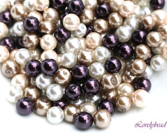 May include: A pile of round, faux pearl beads in three colors: ivory, beige, and dark purple. The beads are scattered on a white surface.