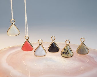 May include: Six triangle-shaped pendants with different colored stones. The stones are white, red, white, black, green and gray. The pendants are on a pink and white agate slice.