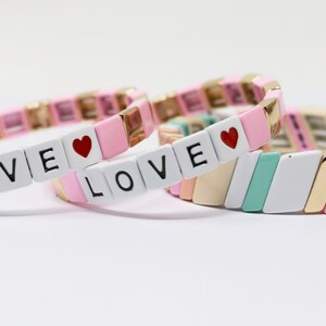 Könnte beinhalten: Drei rosafarbene und weiße Perlenarmbänder mit goldenen Akzenten. Die Armbänder buchstabieren "LOVE" mit einem roten Herz-Anhänger an jedem.