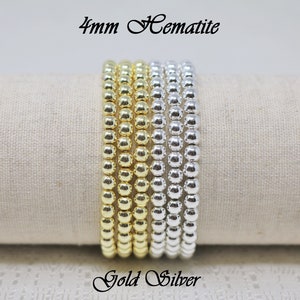 Könnte beinhalten: Gold- und silberfarbene Hämatit-Perlenarmbänder, jeweils mit 4 mm Perlen. Die Armbänder werden auf einem neutralen Stoffhintergrund präsentiert. Der Text auf dem Bild lautet "4mm Hematite" und "Gold Silver".