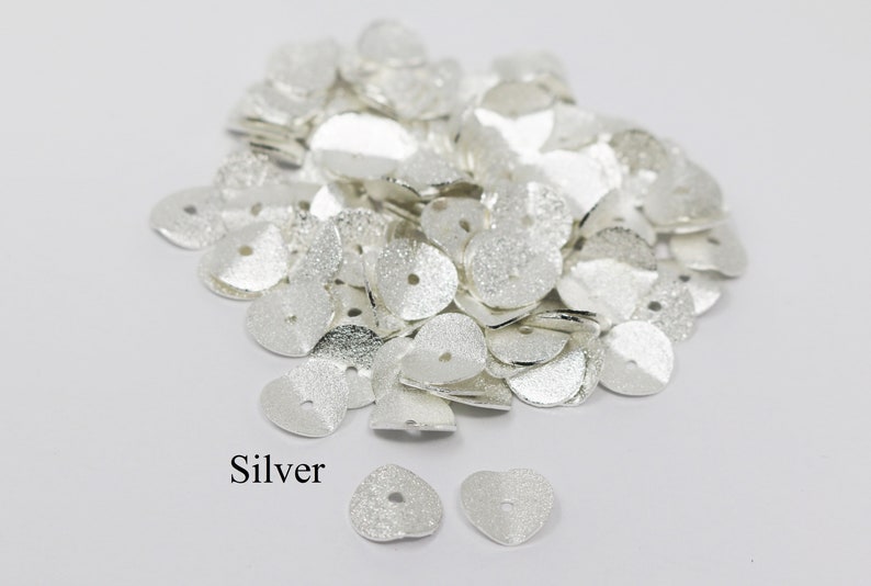 May include: A pile of silver heart-shaped metal beads with a hole in the center. The beads have a textured surface. The word "Silver" is written below the pile of beads.