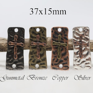 May include: Four metal cross charms in gunmetal, bronze, copper, and silver finishes. Each charm is 37mm x 15mm and has a hammered texture. The charms are displayed on a wooden branch.