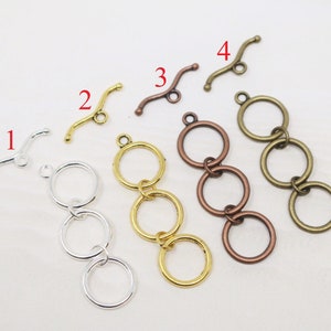 May include: An assortment of jewelry findings, including toggle clasps and triple-ring connectors. The clasps are curved with a loop, and the connectors feature three interconnected rings. Colors include silver, gold, bronze, and copper.