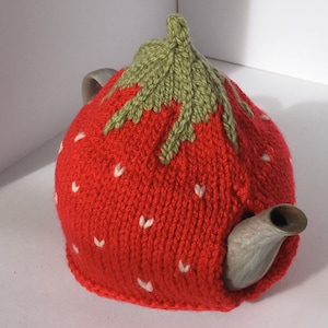 May include: A red knitted teapot cosy shaped like a strawberry with green leaves on top. The cosy is decorated with white embroidered hearts.