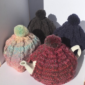 May include: Four knitted teapot cosy covers in different colours. The covers are made with a cable knit pattern and have a pom-pom on top. The colours are pink, grey, blue, and red.
