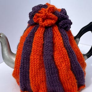 May include: A knitted tea cosy in alternating orange and purple vertical stripes, with a decorative flower on top. The cosy is designed to fit a teapot, providing insulation. The cozy is made of a soft, textured yarn, perfect for keeping tea warm.