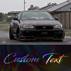 Puede incluir: Un coche negro con una calcomanía de texto personalizado en el parabrisas. La calcomanía es de color arcoíris iridiscente y dice "Custom Text".