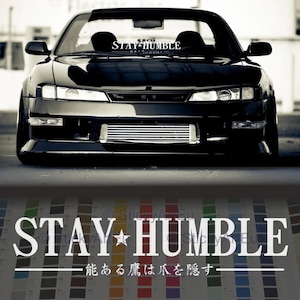 Puede incluir: Un coche negro con una pegatina "STAY HUMBLE" en el parabrisas. El coche tiene una parrilla negra y faros negros. El texto "能ある鷹は爪を隠す" está escrito en blanco sobre un fondo negro debajo del coche.
