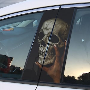 May include: A car window decal featuring a skull with a hand holding it. The skull is detailed with a dark background and a light gray color.
