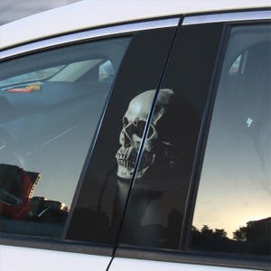 May include: A black and white skull graphic decal for car windows. The skull is detailed and realistic, with a dark background.