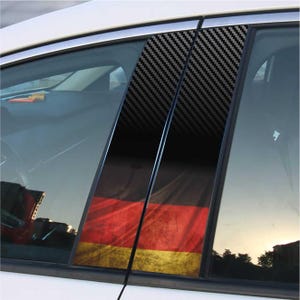 Puede incluir: Calcomanías para ventanas de coche con un patrón de fibra de carbono y la bandera alemana. Las calcomanías son negras, rojas y doradas y se aplican al marco de la ventana del coche. El coche es blanco.