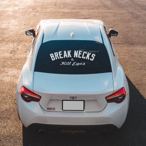 Op de afbeelding: Witte auto met een sticker op de achterruit met de tekst "Break Necks & Kill Egos".