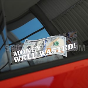 Puede incluir: Una pegatina blanca con el texto "Money Well Wasted!" sobre un fondo de billete de 100 $. La pegatina está en la ventana de un coche rojo.