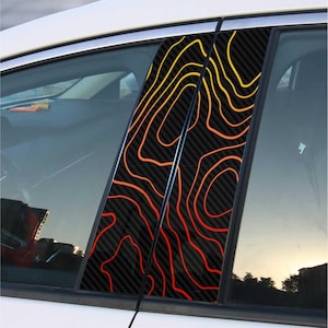 May include: Car window pillar decals with a topographic map design. The decals feature a black carbon fiber background with orange and yellow contour lines. The decals are applied to the car's window pillars.