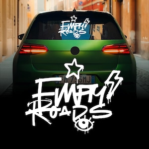 Pode incluir: Um carro verde com o texto "EMPTY ROADS" em letras brancas estilo grafite no vidro traseiro e no para-choques. Um desenho de estrela e raio também está incluído. O carro está estacionado numa rua estreita.