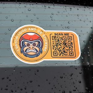 Könnte beinhalten: Ein orangefarbener Aufkleber mit einem Comic-Gorilla, der einen Helm und eine Sonnenbrille trägt. Der Aufkleber trägt die Aufschrift "CUSTOM LOGO AND QR STICKERS" und "SCAN ME" mit einem QR-Code. Der Aufkleber befindet sich auf einer nassen, dunklen Oberfläche.